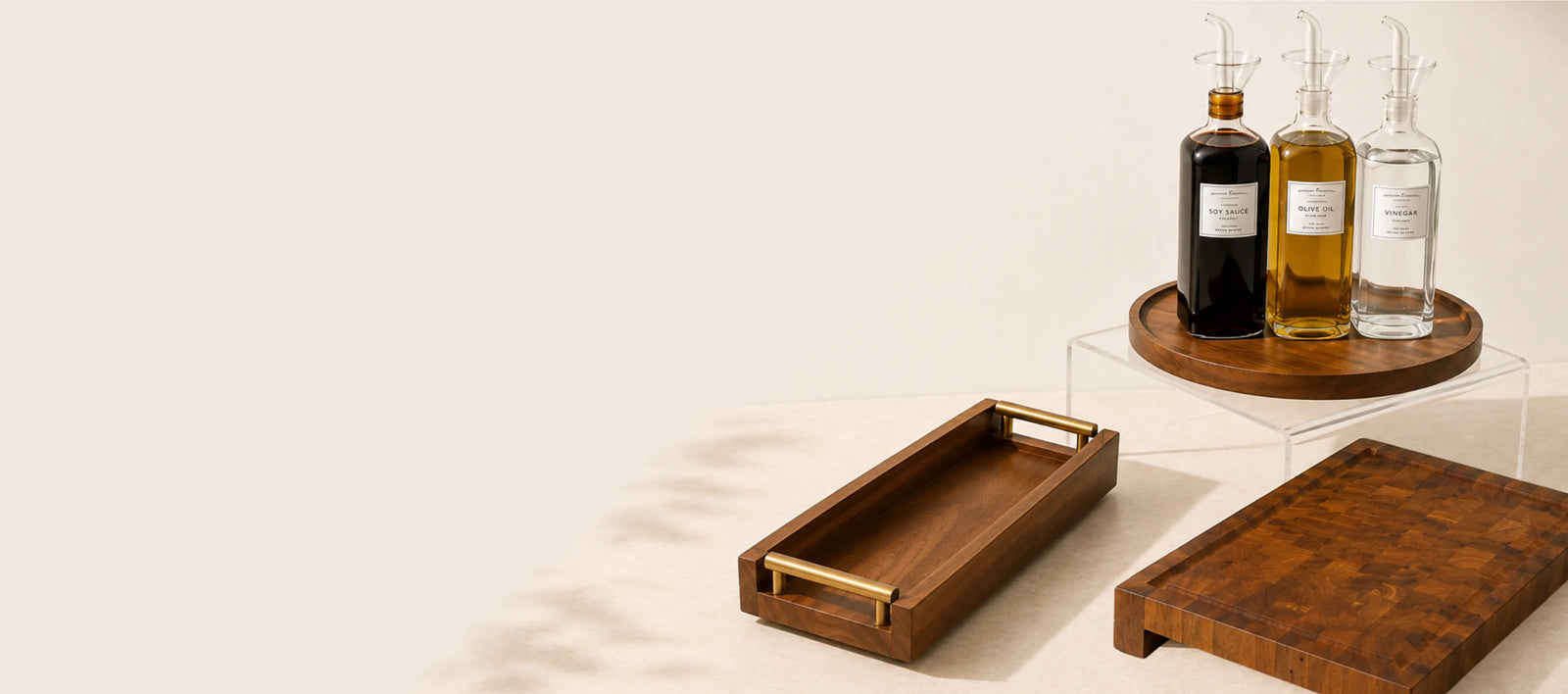 A styled product shot featuring a rectangular wooden serving tray with brass handles in the foreground, a matching wooden cutting board, and a round wooden tray holding three glass bottles on a clear acrylic riser in the background. The scene is set against a warm beige background with soft leaf shadows.